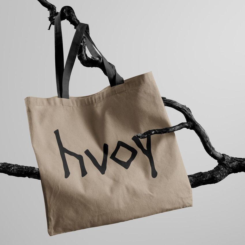 Tote bag mockup featuring a beige fabric bag with black handles, hanging on a dark, textured tree branch against a light gray background.