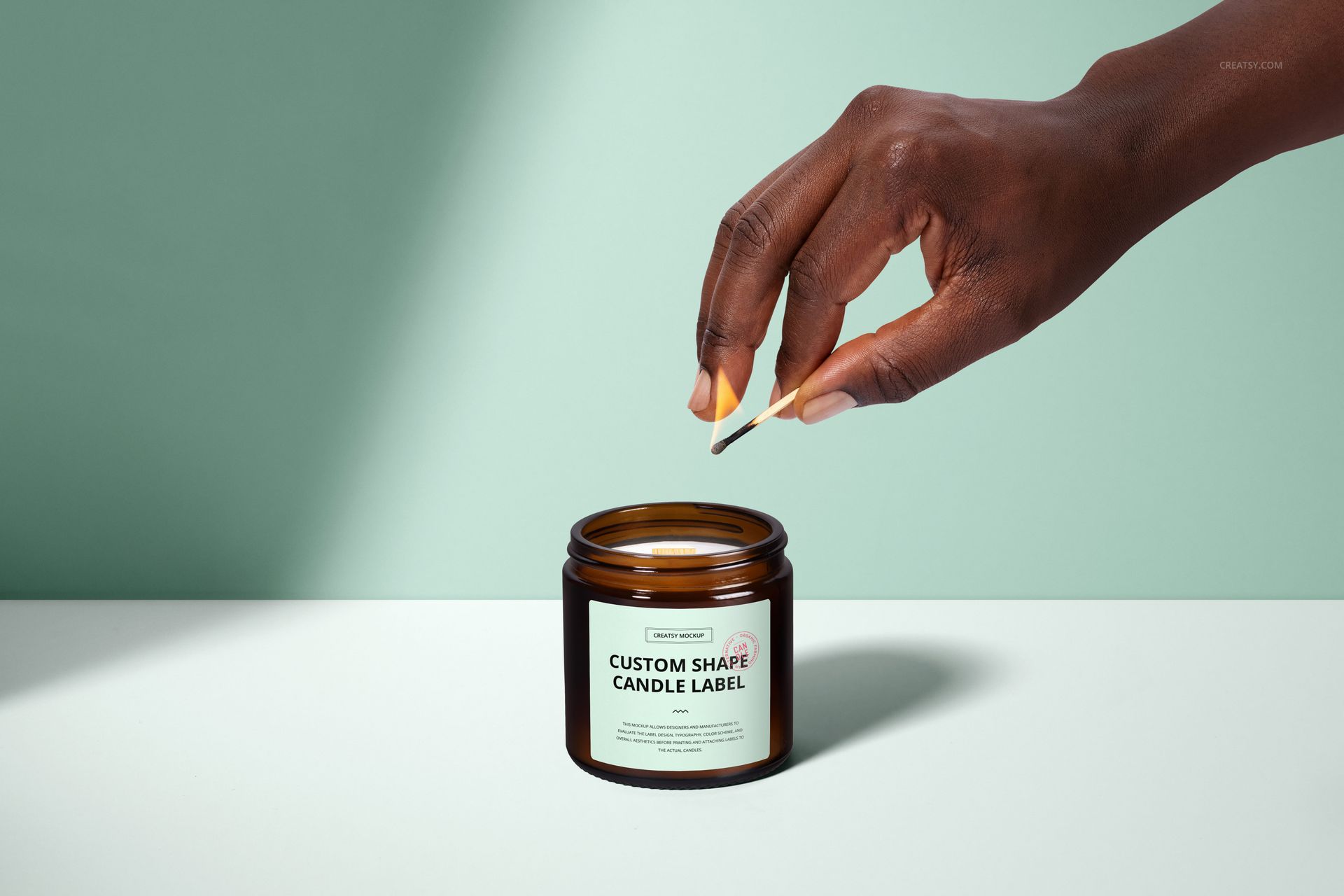 Amber glass jar candle with wooden wick on a white surface, hand lighting the candle, front view mockup scene.