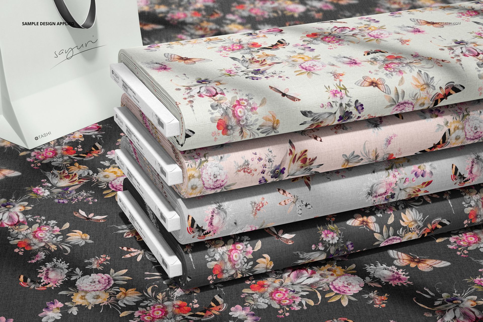 Fabric bolts mockup showing layered textiles with botanical designs in light and dark tones, viewed from a side angle.