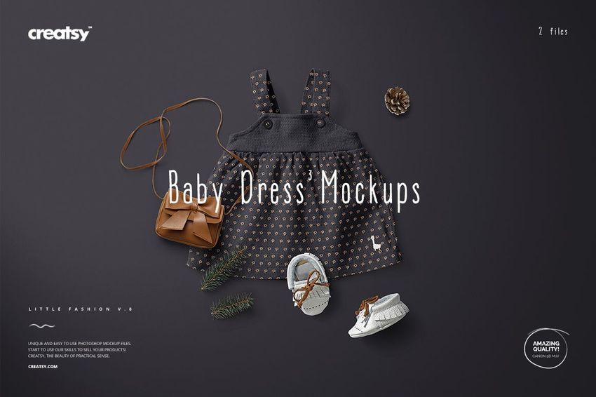 Front view of a baby dress mockup with a dark patterned fabric, displayed alongside baby shoes, a small purse, and pinecones.