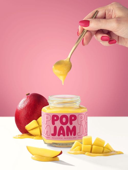 Front view of a mango jam jar mockup with a pink label, surrounded by fresh mango slices and a hand holding a spoon.