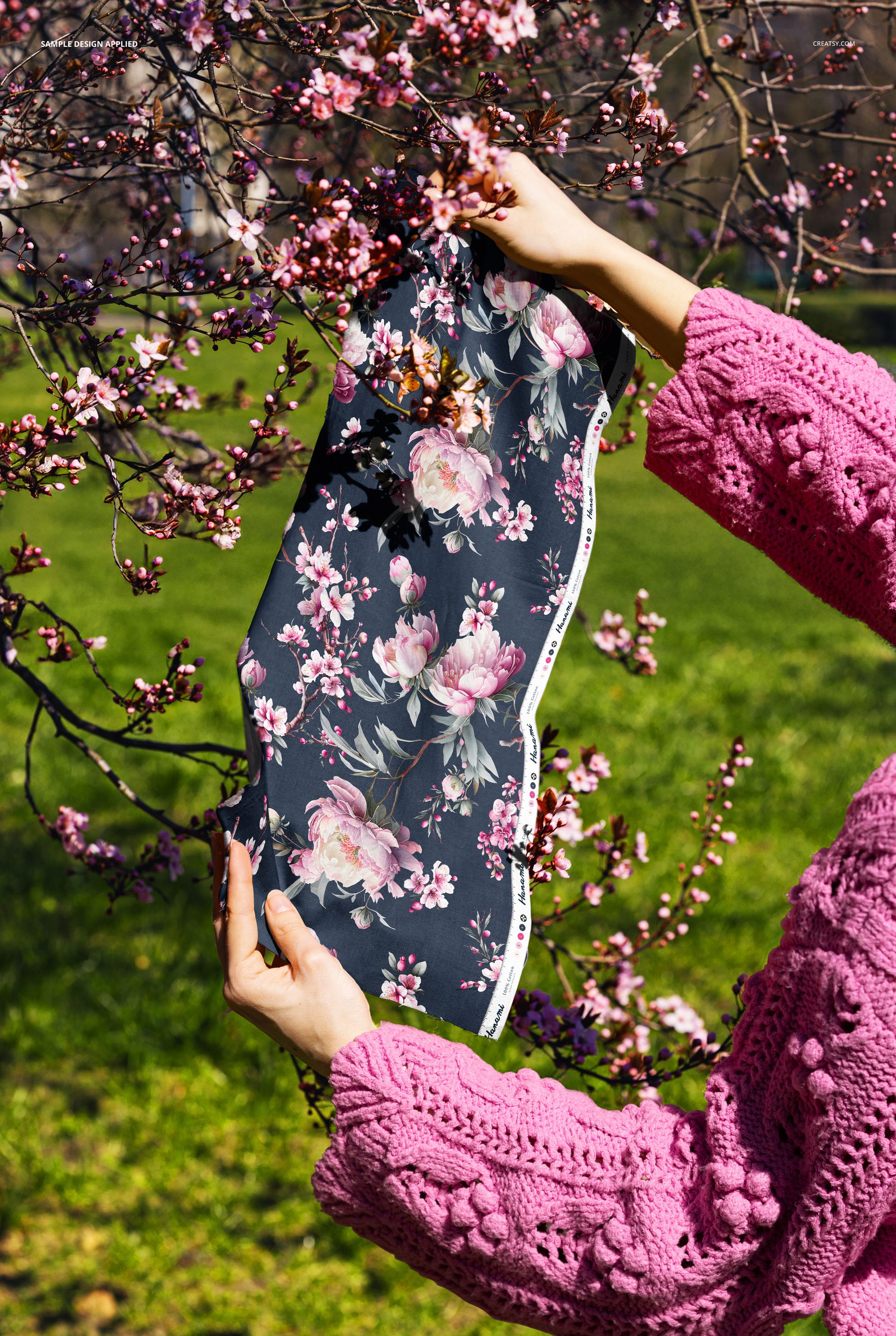 Floral fabric swatch mockup shown outdoors, held by hands wearing a pink textured sweater near blossoming tree branches.