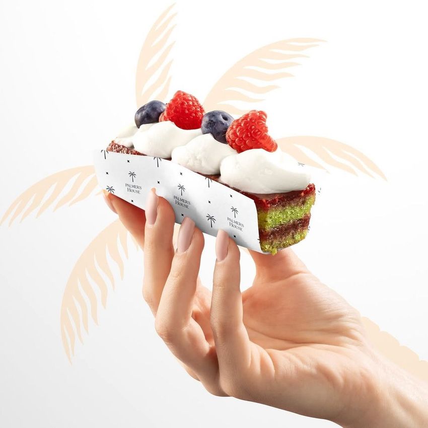Hand holding a dessert wrapped in food safe paper with a palm tree pattern, topped with whipped cream, raspberries, and blueberries.