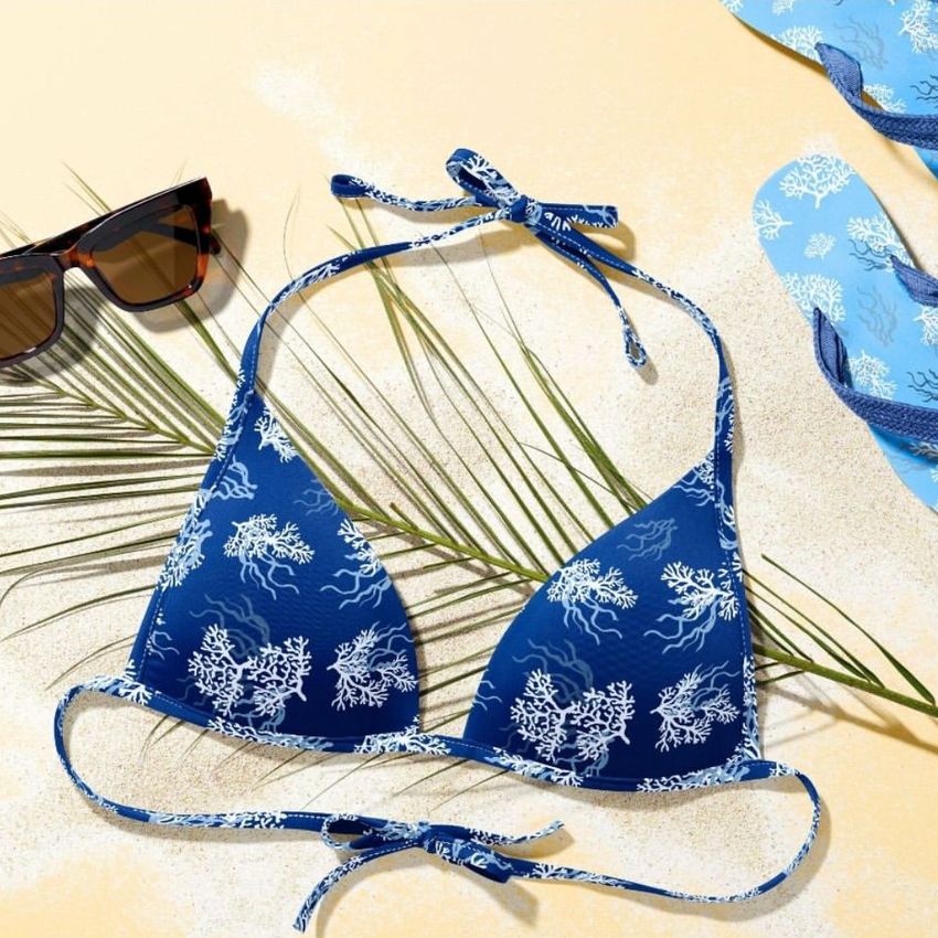 Bikini mockup featuring a blue bikini with white coral patterns, laid flat on sand with sunglasses, palm leaves, and flip-flops.