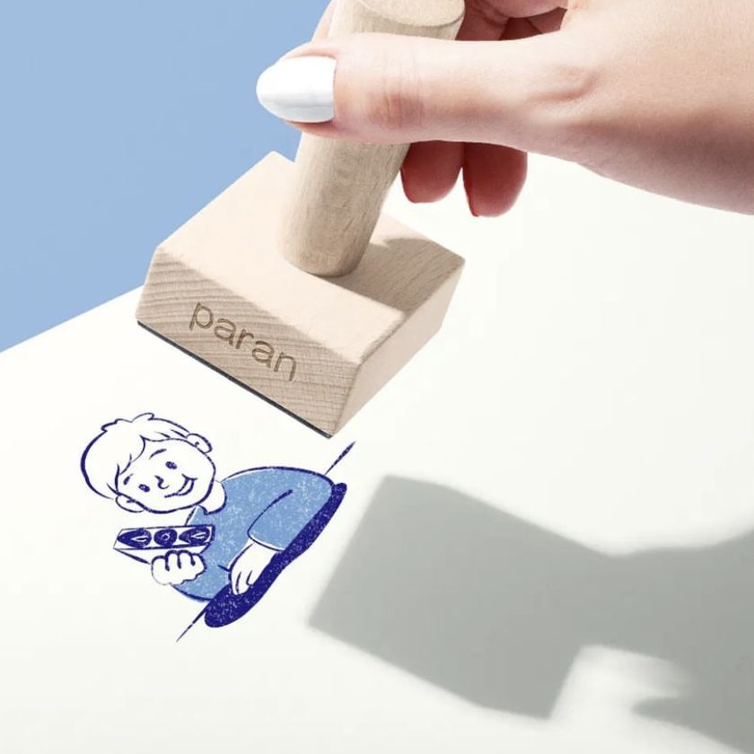 Person's hand using a wooden stamp mockup on a light surface, leaving a blue ink image of a cartoon figure with short hair.
