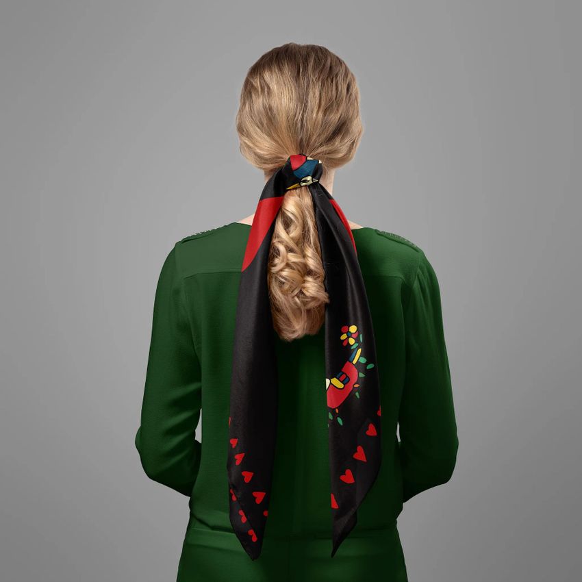 Silk scarf mockup showing a woman from behind with blonde hair tied in a ponytail, wearing a green blouse and a black scarf with red and colorful abstract patterns.