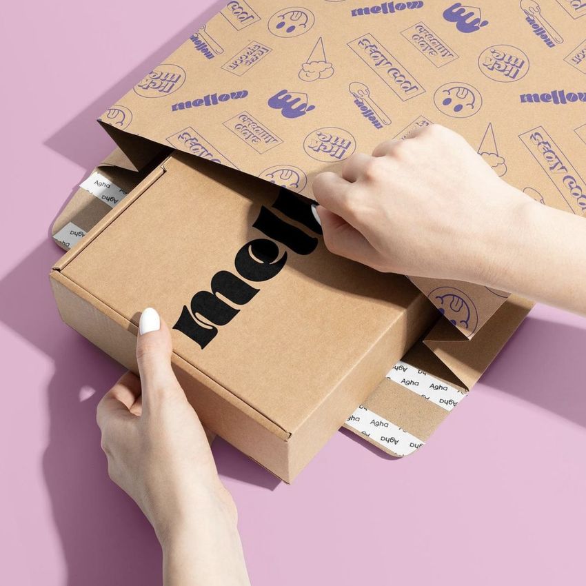 Person assembling a kraft mailer mockup with cartoon-style graphics and a brown box, set against a light pink backdrop.
