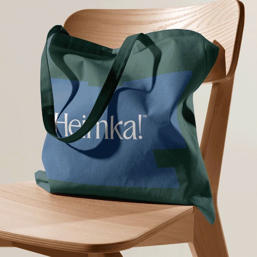 Tote bag mockup featuring a green and blue fabric bag with a bold text overlay, draped over a light wooden chair.