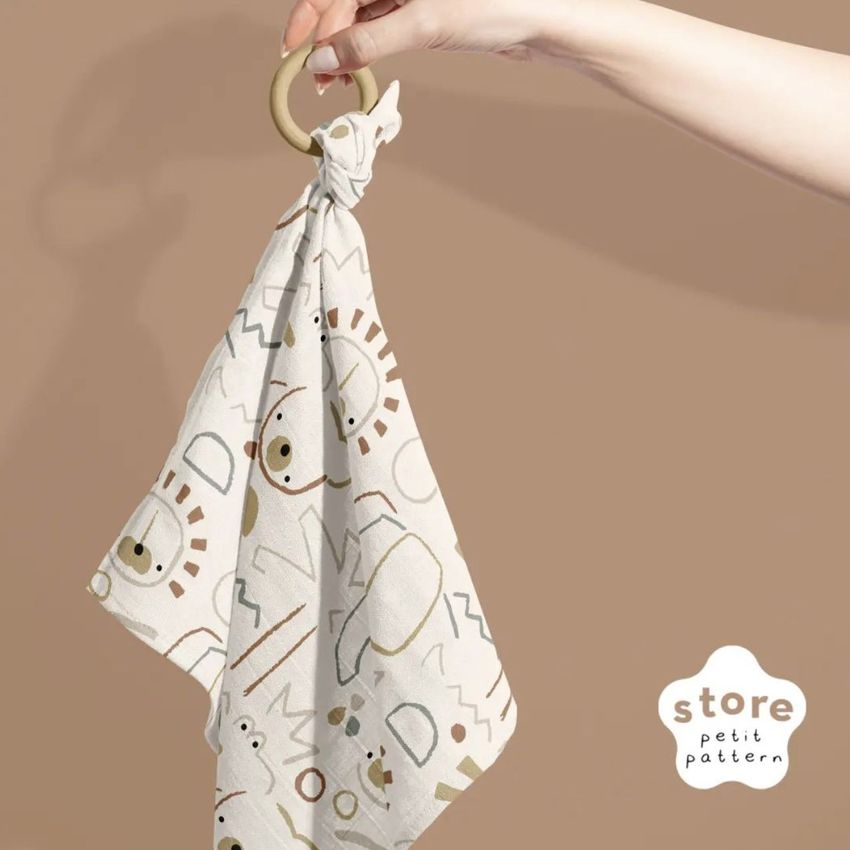 Muslin cloth teether mockup with abstract animal faces and shapes, held by a hand against a solid brown backdrop.