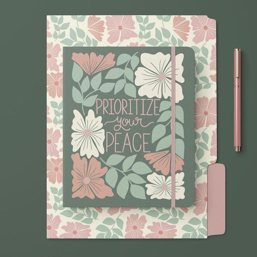 Stationery set mockup featuring a floral-patterned folder, matching notebook with text overlay, and a pink pen on a green surface.