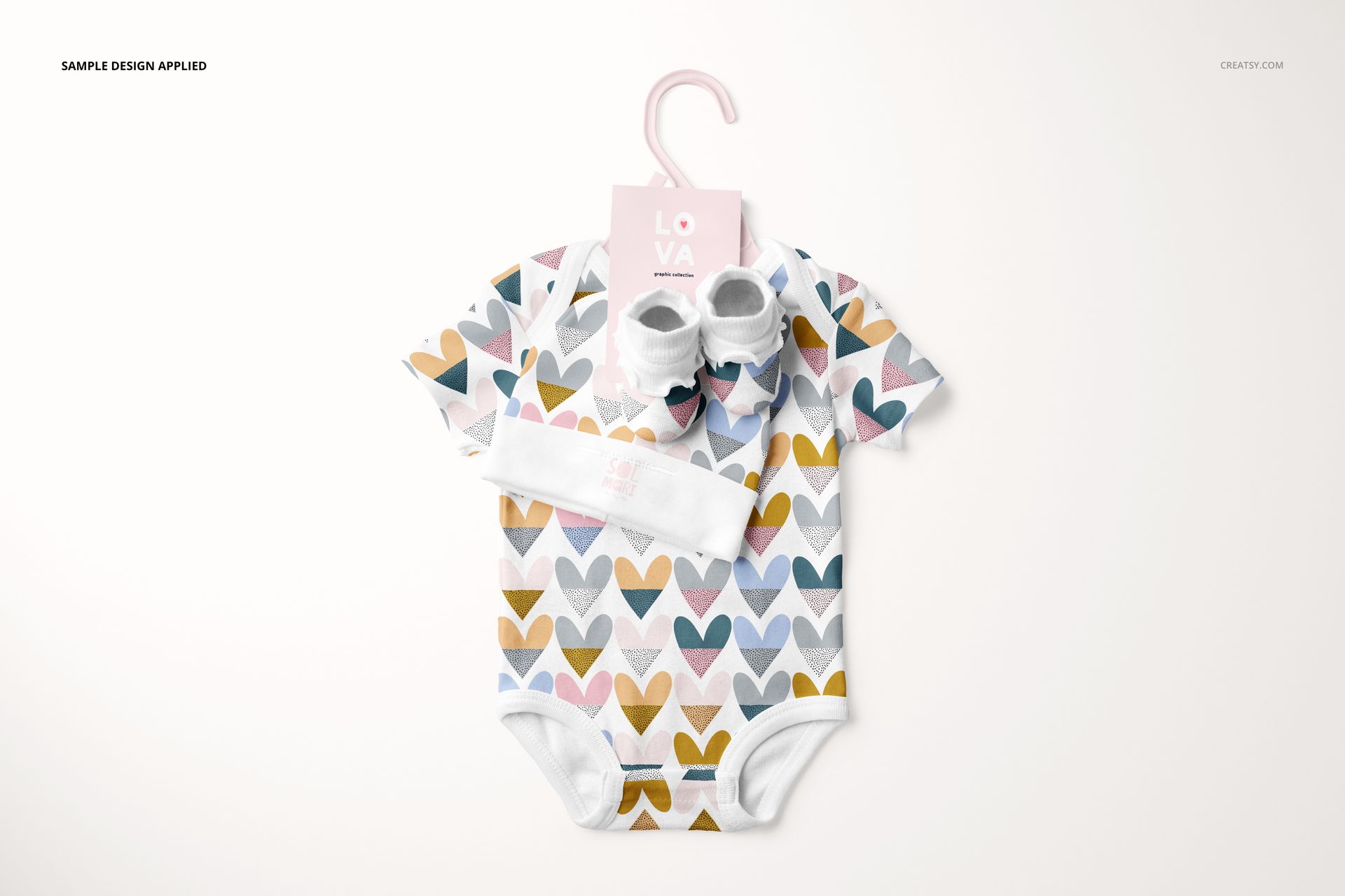 Front view of a baby onesie mockup with pastel heart print, accompanied by booties and hat, set on a white surface.