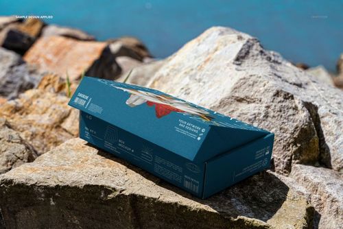 Blue mailing box mockup on uneven rocks by the sea, showing side and top panels with printed graphics.