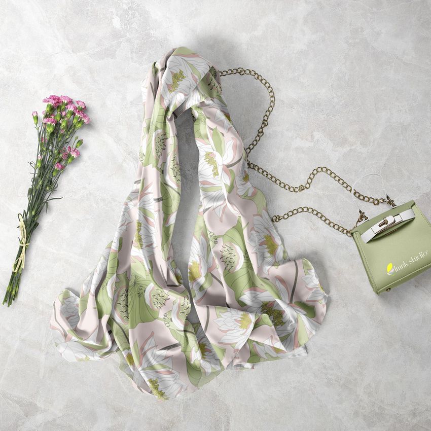 Chiffon silk scarf mockup loosely laid on a light stone surface, featuring a floral pattern in green and white tones, with a small green purse and a bouquet of pink flowers nearby.
