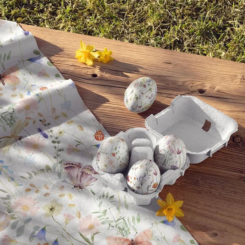 Cotton fabric with a wildflower pattern is draped beside a carton of decorated Easter eggs on a wooden surface outdoors.