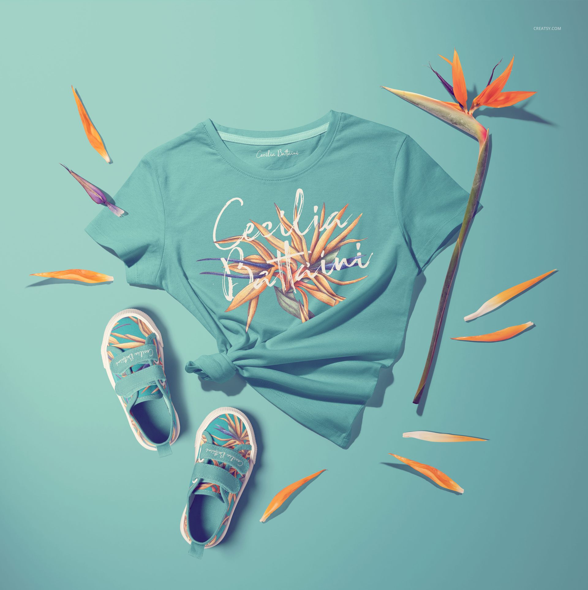 Top view of a kids T-shirt mockup set with shoes and tropical flowers, displayed on a pastel blue background.