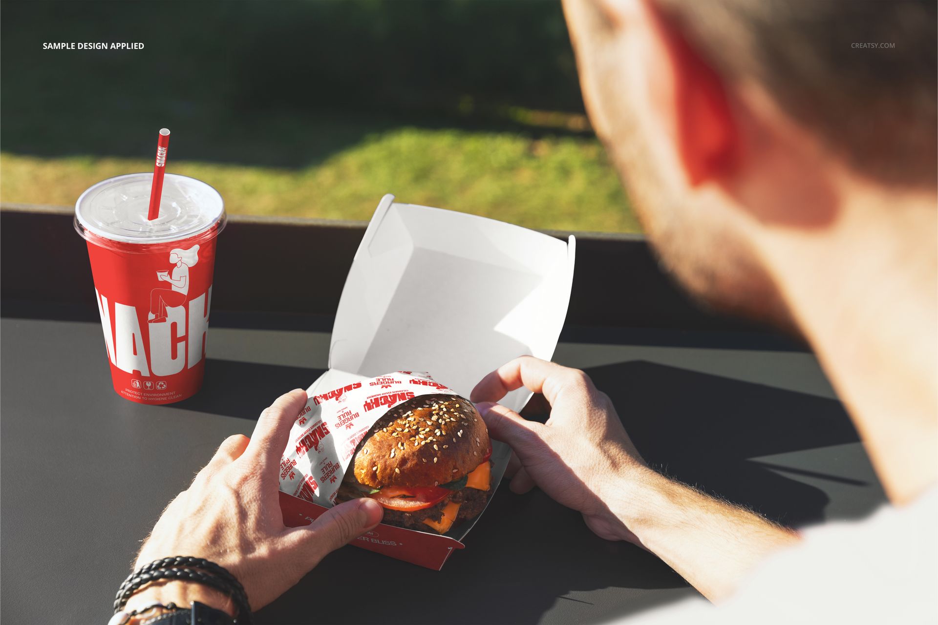 Person holding a burger wrapped in branded paper with a drink cup on a table, outdoor setting, front view mockup.