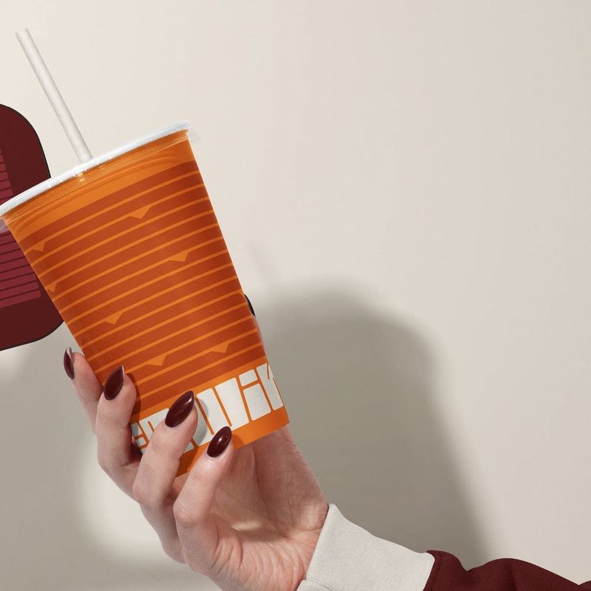 Disposable cup mockup featuring orange horizontal stripes and abstract shapes, held by a hand with dark nail polish.