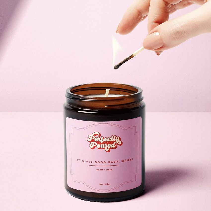 A hand lights a brown glass candle jar with a pink sticker label on a pastel pink background, shown in a sticker mockup.