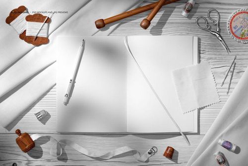 Top view of a creative desk with white cotton fabrics, open blank notebook, pen, scissors, and sewing props.