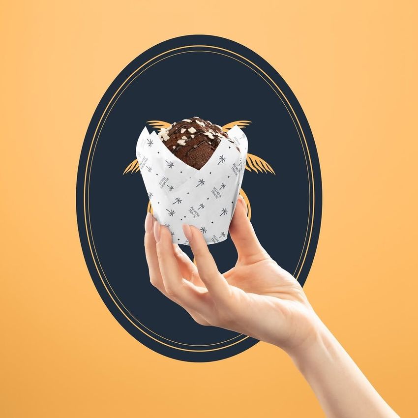 Hand holding a chocolate muffin wrapped in patterned food safe paper, with a decorative oval design on a peach background.