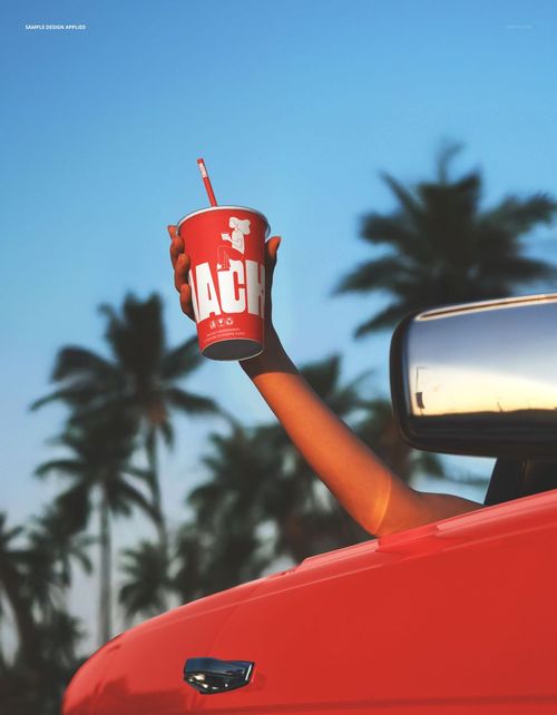 PSD mockup showing a red paper beverage cup with straw held from a car window, sunny outdoor scene with palm trees.