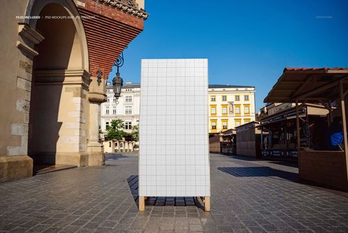 Town square signboard mockup with a blank grid pattern, standing upright on a stone pavement with historic buildings in the background.
