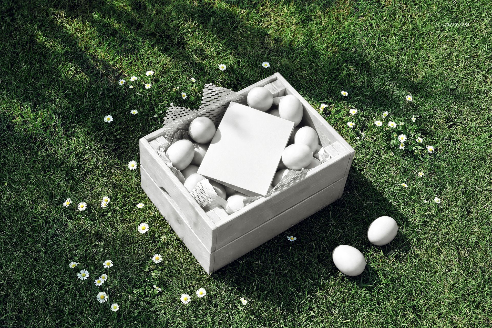 PSD mockup featuring a wooden crate filled with Easter eggs and a blank card, placed on grassy lawn with daisies.