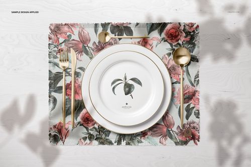 PSD mockup showing a floral fringed placemat, decorative white plate, and gold utensils arranged on a light surface.
