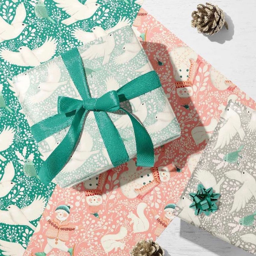 Gift box wrapping paper mockup featuring pastel green and pink sheets with snow people, birds, and foliage patterns, seen from above.