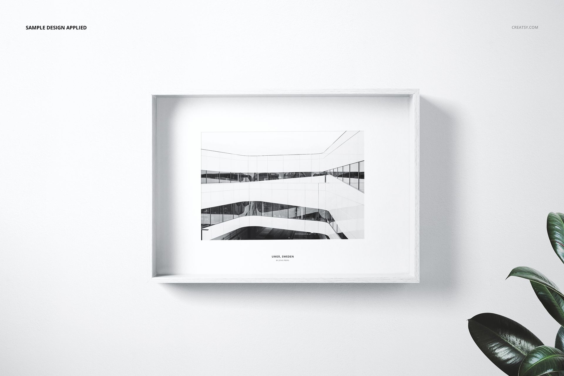 Front view of a white wooden frame mockup displaying a black and white architectural artwork, with a plant nearby.