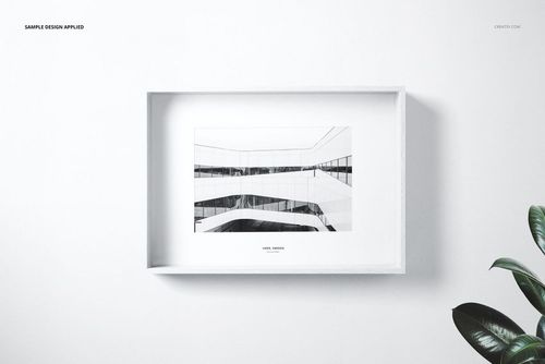 Front view of a white wooden frame mockup displaying a black and white architectural artwork, with a plant nearby.