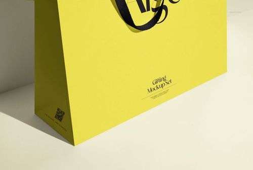 Yellow paper gift bag mockup with black handles, photographed from a side angle on a neutral background.