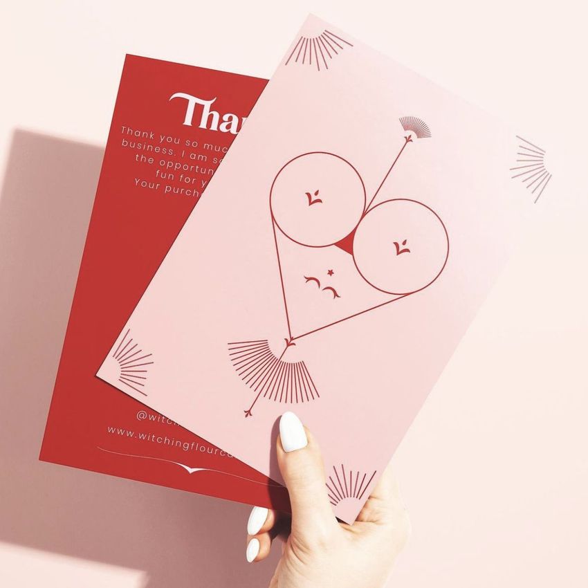 Hand holding two stationery cards, one pink with a minimalist face illustration and one red card with white text, on a light background.