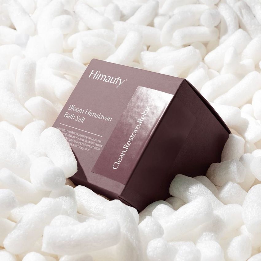 Square box packaging mockup with a dark mauve design and white text, surrounded by white packaging peanuts in bright lighting.
