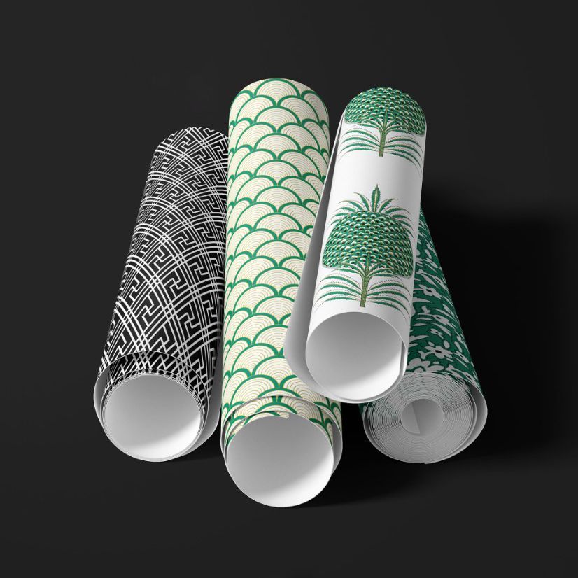 Paper wallpaper mockup featuring four rolled sheets with geometric and botanical patterns in green, black, and white on a dark surface.