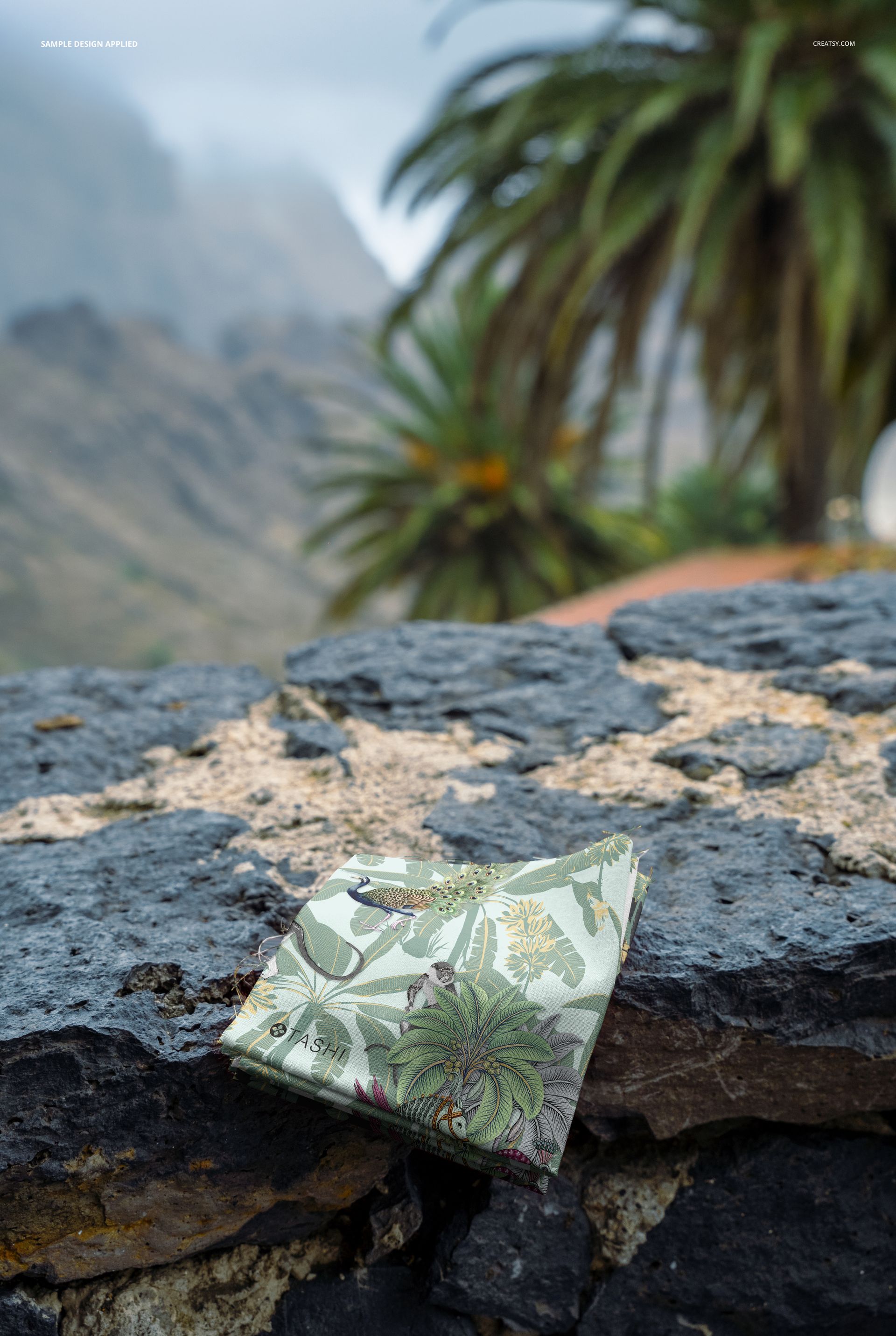 PSD mockup showing a pile of decorative fabrics placed on textured volcanic rock, surrounded by tropical greenery and distant peaks.
