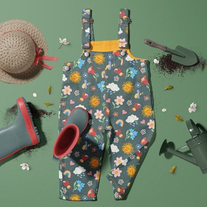 Kids dungarees mockup featuring colorful garden-themed patterns, laid flat on a green surface with gardening props around.