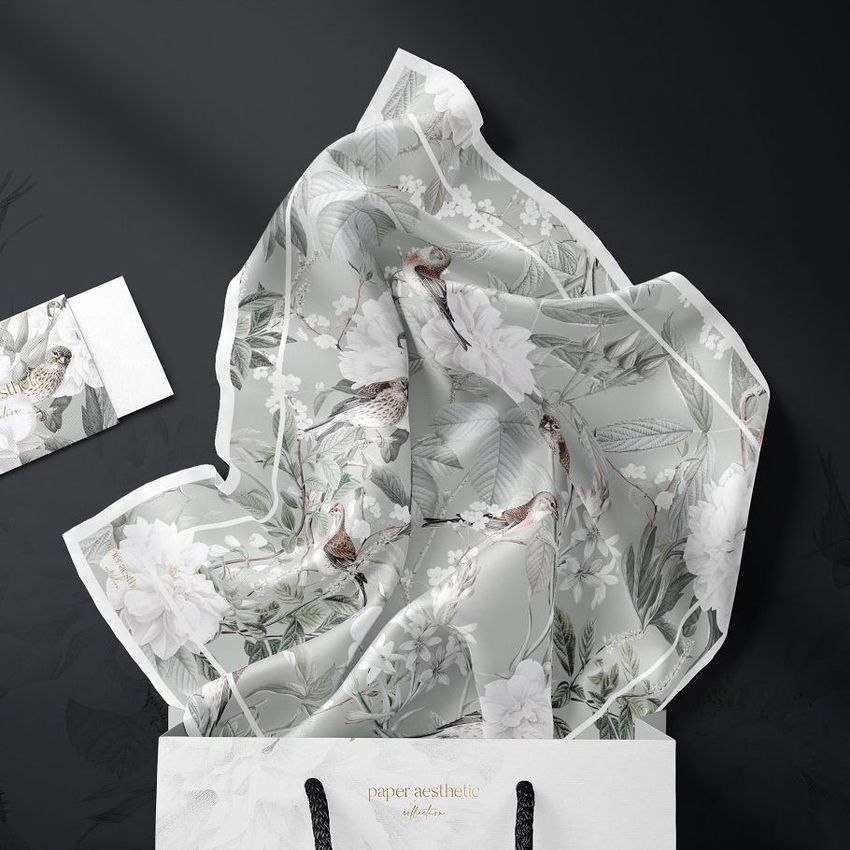 Silk scarf mockup featuring a botanical pattern with birds and flowers, draped over the edge of a white shopping bag on a dark surface.