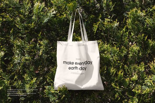 White tote bag with black text hanging on green bush, front view, natural outdoor lighting, PSD mockup.