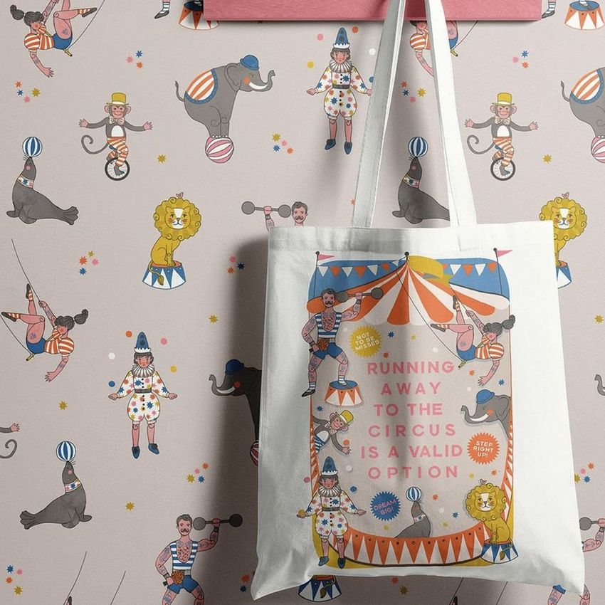 Tote bag mockup featuring a circus-themed design with colorful animals and performers, hanging against a matching patterned wall.