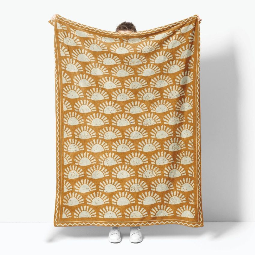 Person holding a fleece throw blanket mockup with a repeating cream sunburst pattern on a mustard yellow background against a white wall.
