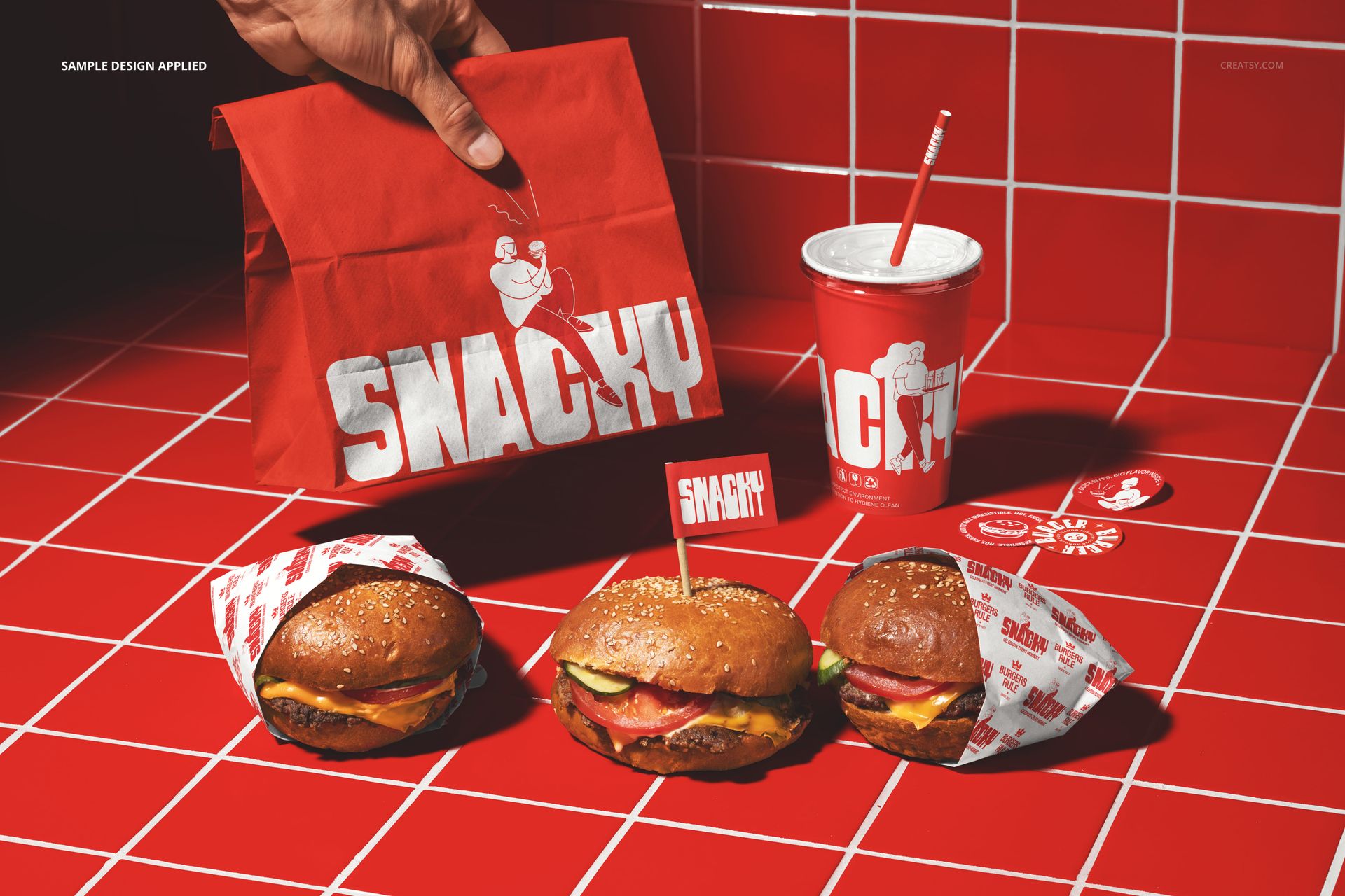 Front view of a take-out burger set mockup featuring three burgers, a paper bag, a drink cup, and red tiled background.