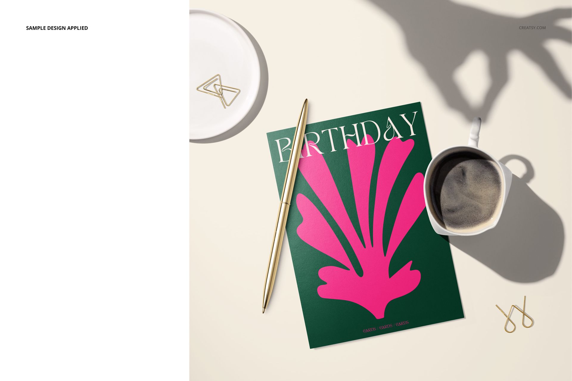 Top view of a 5x7 greeting card mockup featuring a green card with pink artwork, gold pen, coffee, and paper clips.