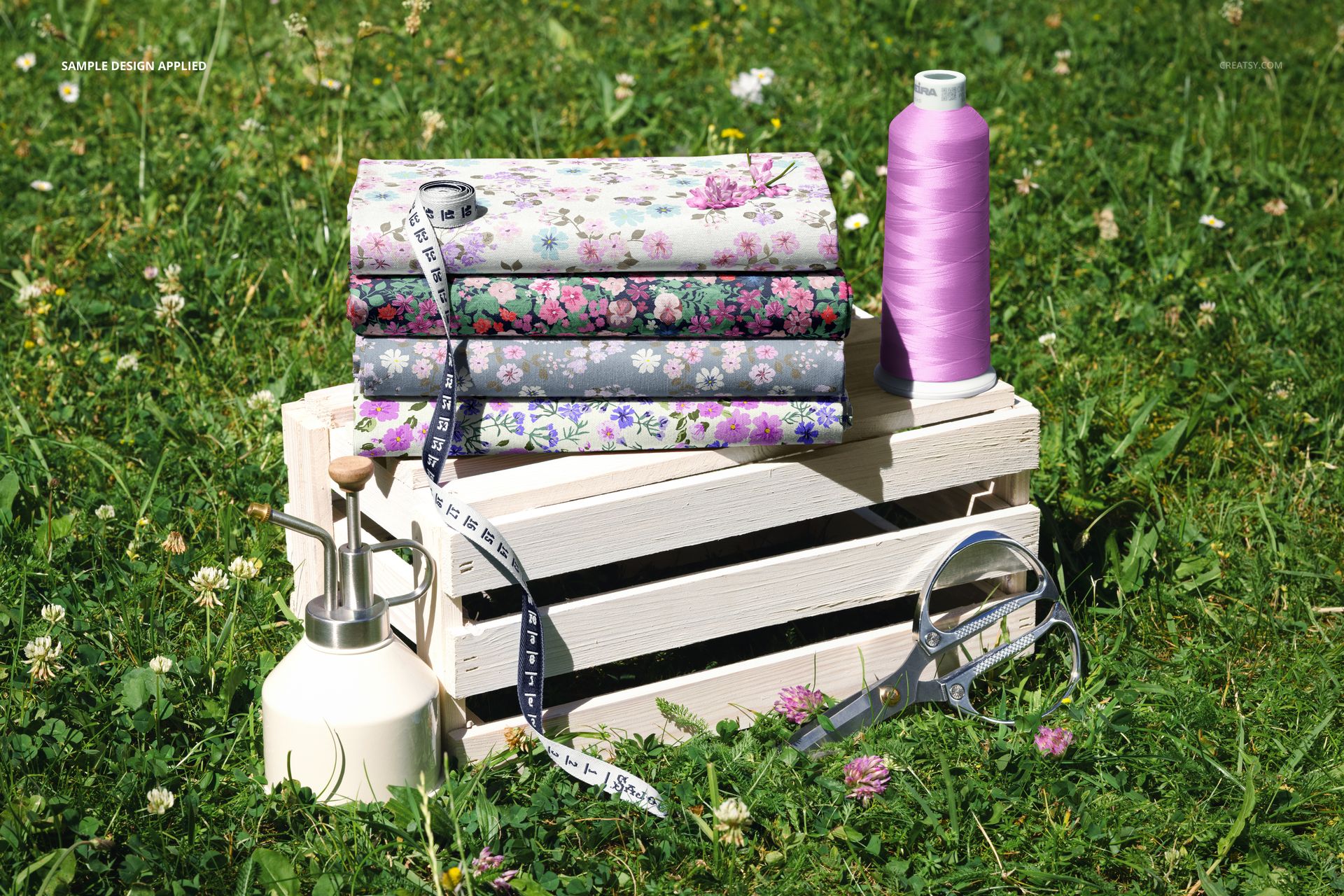 PSD mockup featuring a pile of patterned jersey fabric on a crate in a grassy area, with sewing accessories visible.