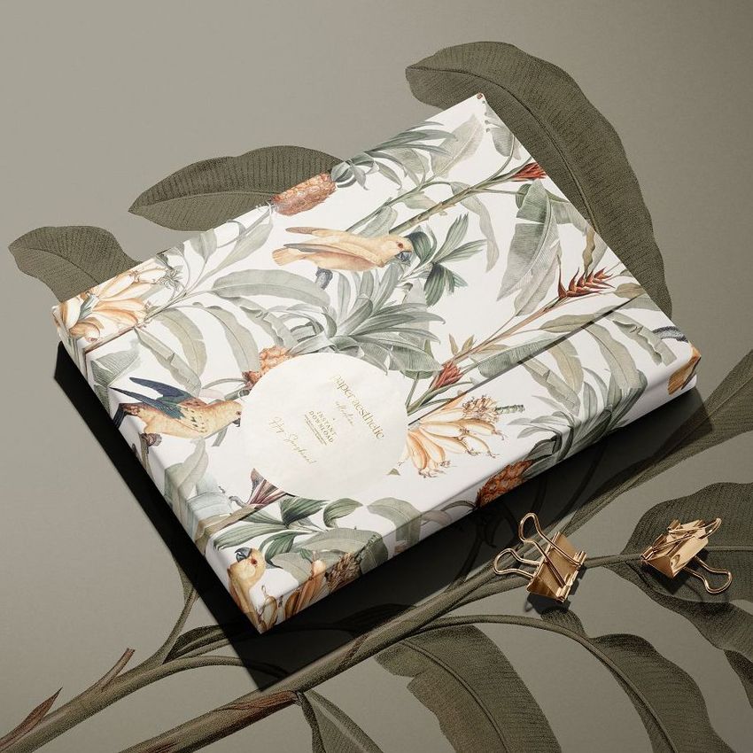 Sticker mockup featuring a rectangular package wrapped in tropical patterned paper with birds and leaves on a neutral surface.