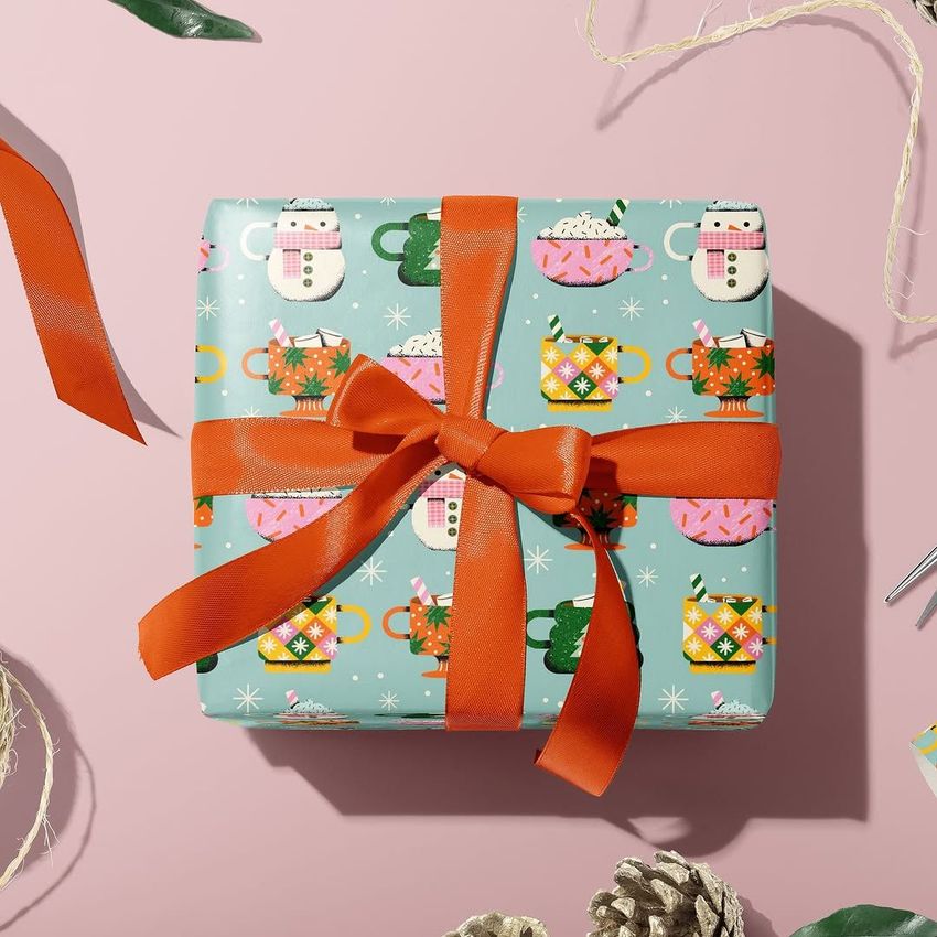Wrapping paper mockup featuring a pastel blue background with hot cocoa mugs and whipped cream patterns, tied with a large orange ribbon.