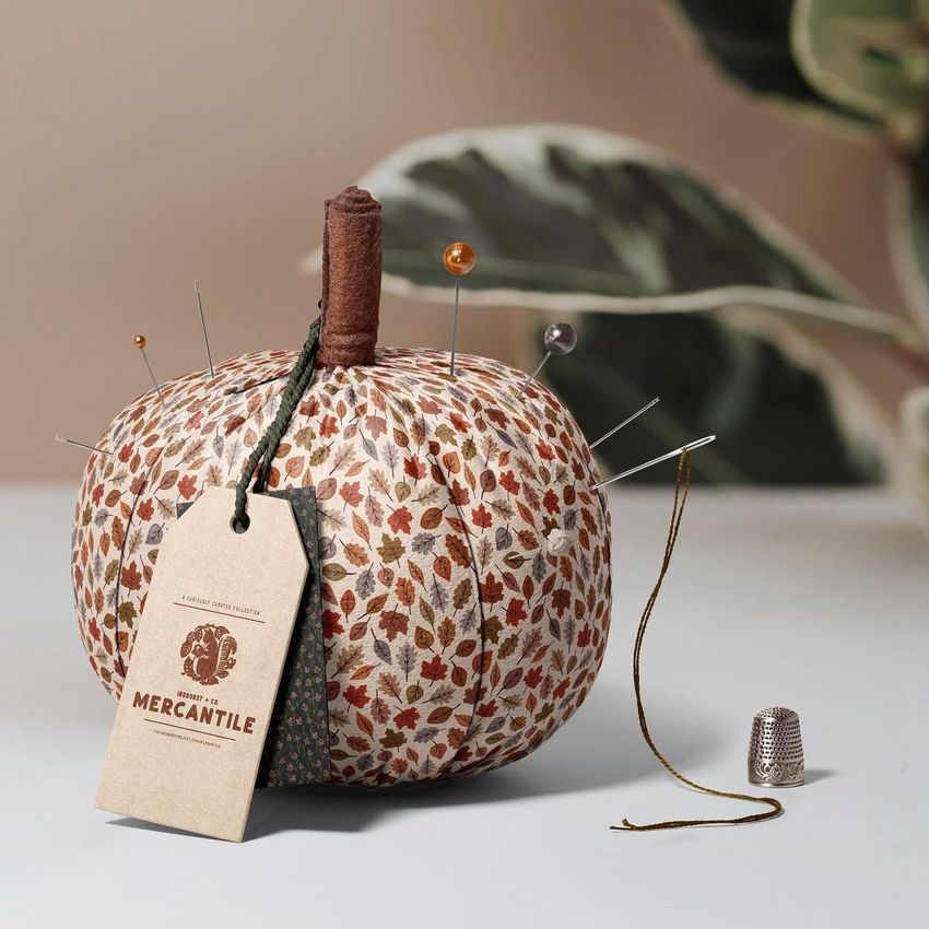 Fabric pumpkin mockup with an autumn leaf pattern, used as a pincushion with pins, a tag, and a thimble on a neutral surface.