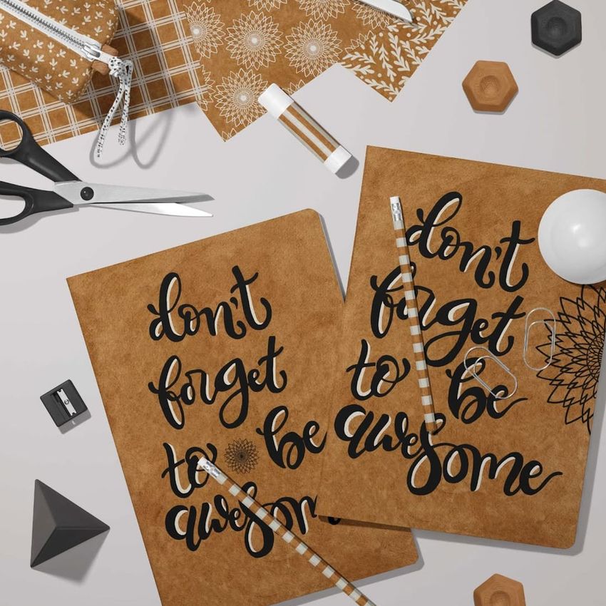 Stationery mockup featuring two classic A4 notebooks with brown covers, bold black script text, and geometric props on a light surface.