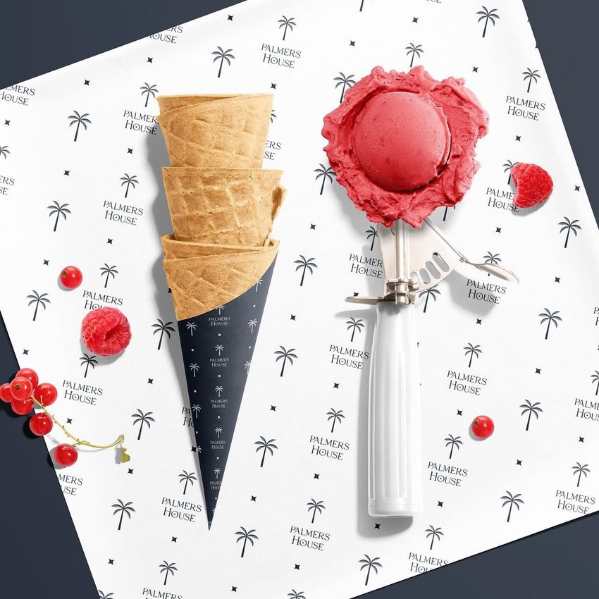 Ice cream paper mockup featuring a black cone wrapper and white sheet with palm tree and text pattern, alongside a metal scoop with pink ice cream.