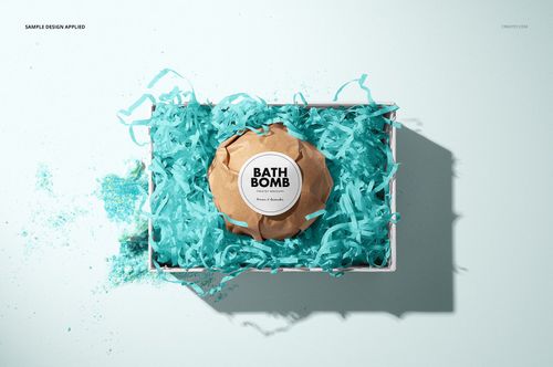Bath bomb mockup in a rectangular box filled with blue paper shreds, viewed from above on a light background.